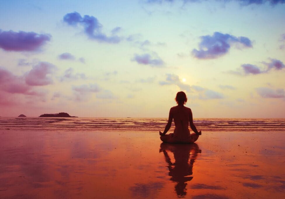 Person meditating on beach during sunset.